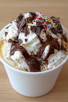 Creamy vanilla ice cream topped with whipped cream, chocolate sauce drizzle, chocolate shavings and colorful sprinkles served in white paper cup. Sweet dessert treat.