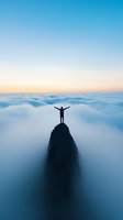 Silhouette of man standing with raised arms on mountain peak above clouds at sunrise, expressing freedom and achievement in breathtaking aerial landscape.
