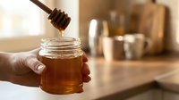 Closeup of hand holding glass jar while pouring golden honey with wooden dipper in bright kitchen setting, ideal for food and wellness content.