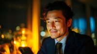 Professional Asian businessman in dark suit using smartphone in modern office with warm evening lighting and city bokeh background for corporate communications.