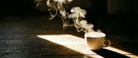 Steaming white cup of hot coffee or tea on a rustic wooden surface with dramatic light and rising smoke in a dark moody setting.
