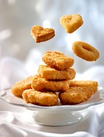 Golden crispy chicken nuggets stacked on white plate with heart-shaped pieces floating in air against soft light background.