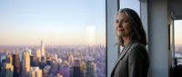 Professional businesswoman with long gray hair gazing thoughtfully out office window at city skyline during golden hour, representing success and leadership.