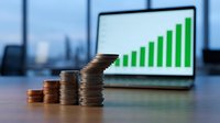 Stacked coins arranged in ascending order with upward trending green bar chart on laptop screen, symbolizing financial growth, investment returns, and business success metrics.