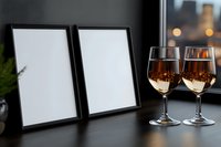 Two empty black photo frames and wine glasses filled with rose wine on windowsill against blurred city lights background during evening hours.