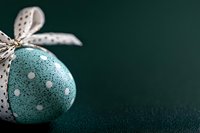 Speckled turquoise Easter egg decorated with white polka dots and tied with spotted ribbon on dark green background, festive spring holiday composition.