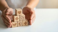 Hands protecting wooden blocks with healthcare and medical symbols including heart, cross, shield and stethoscope representing health insurance coverage.