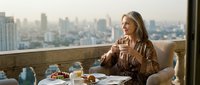Woman in brown jacket holding coffee cup on high-rise balcony overlooking cityscape. Breakfast setting with pastries and beverages. Urban lifestyle and morning routine concept for lifestyle, hospitality, and travel content.