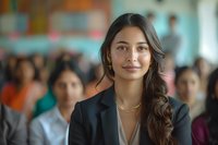 Young Asian businesswoman with confident smile wearing black blazer at corporate conference or meeting, with blurred audience in background. Professional workplace setting.