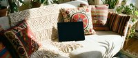 Cozy bohemian living room interior with laptop on white sofa surrounded by colorful ethnic pillows and houseplants for remote work and relaxation.
