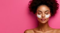 Young African American woman with curly hair applying white face mask cream on pink background for skincare routine and beauty treatment.