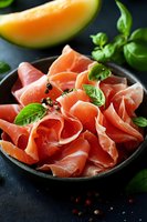 Sliced prosciutto ham with fresh basil leaves and cantaloupe melon on dark plate. Gourmet Italian appetizer with herbs and peppercorns for culinary presentations.