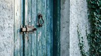 Weathered blue wooden door with rusty metal latch and ring handle on textured white stone wall. Vintage Mediterranean architecture detail with peeling paint.