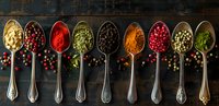 Colorful spices and peppercorns in vintage silver spoons arranged in row on dark rustic wooden background, scattered seasoning ingredients for cooking.