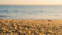 Sandy beach shore with blurred ocean and sky in background, shallow depth of field focusing on golden sand grains and pebbles, suitable for travel and nature backgrounds.