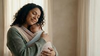 Young mother with curly hair holding newborn baby in carrier wrap at home, peaceful bonding moment between parent and infant in soft natural light.