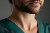Male healthcare professional with dark beard wearing teal scrubs, cropped portrait showing neck and shoulders against neutral background for medical, wellness, and professional use.