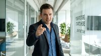 Confident Caucasian businessman in dark suit pointing finger at camera in modern glass office corridor with motivational text on wall.