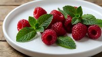 Fresh red raspberries with mint leaves arranged on white plate, perfect for food photography, healthy eating, dessert, and culinary content.
