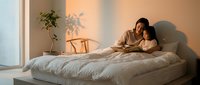 Young Asian couple embracing intimately on bed in warm sunlit bedroom with minimalist decor and plant shadows on wall creating cozy romantic atmosphere.