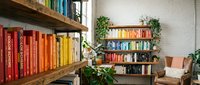 Cozy home library with colorful books organized by rainbow spectrum on wooden shelves, plants and reading chair creating warm literary atmosphere.