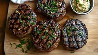 Grilled beef tenderloin steaks with fresh herbs and garlic butter sauce on wooden cutting board, charred grill marks and juicy medium rare cooking.