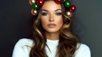 Young woman with long wavy brown hair wearing white sweater and Christmas ornament wreath headpiece with red and green baubles against dark background. Holiday beauty portrait.