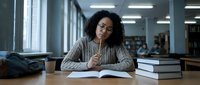 Young African American woman student studying in modern university library with books and notebooks, focused on academic research and learning.