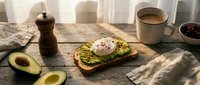 Avocado toast with poached egg on rustic wooden table, coffee mug and fresh avocado halves in warm morning light.