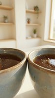 Two ceramic bowls filled with dark liquid on wooden surface in bright modern kitchen with floating shelves and potted plants creating cozy morning atmosphere.