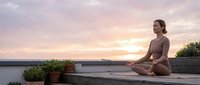 Young woman practicing meditation yoga pose on wooden deck during golden hour sunset with ocean view and potted plants creating peaceful wellness atmosphere.