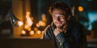 Young Caucasian man smiling while working on laptop at night, warm bokeh lights in background creating cozy atmosphere for remote work or online communication.