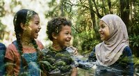 Three diverse young women laughing together outdoors in natural forest setting with sunlight filtering through trees creating joyful friendship moment.