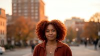 Young African American woman with curly hair smiling outdoors in urban setting during golden hour sunset with warm autumn lighting and city buildings.
