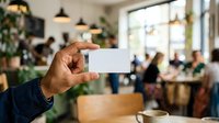 Man holding blank white business card in modern cafe with plants and blurred background, suitable for branding, networking, and corporate identity mockups.