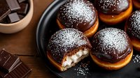 Sweet donuts filled with cream, topped with dark chocolate glaze and powdered sugar on black plate. One donut cut to show creamy filling. Dark chocolate pieces aside.
