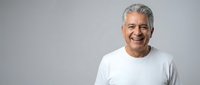 Cheerful middle-aged Latino man with gray hair smiling confidently, wearing a white t-shirt, isolated on a light gray background with copy space.