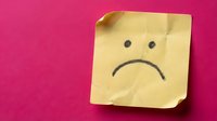 Yellow sticky note with sad face drawn in black ink on bright pink background expressing negative emotions and disappointment.
