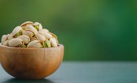 Roasted pistachios in wooden bowl against green blurred background, healthy nuts and snacks for vegetarian nutrition and natural food photography.
