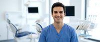 Smiling male dentist in blue scrubs standing in modern dental clinic with equipment and chair visible in background for healthcare and medical practice concepts.
