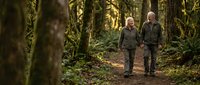 Senior couple walking together on forest trail surrounded by moss-covered trees and lush green vegetation enjoying peaceful nature hike in woodland setting.