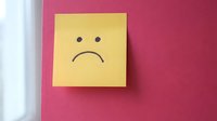 Yellow sticky note with sad face drawn on pink background expressing negative emotions and disappointment for mental health awareness campaigns.
