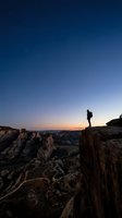 Lone hiker silhouette standing on rocky cliff edge at twilight with starry sky and canyon landscape below for adventure and exploration concepts.