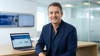 Confident middle-aged Caucasian businessman in navy suit smiling at modern office desk with laptop displaying financial charts and data analytics.
