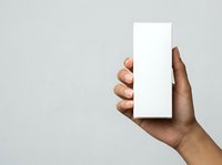 Male hand with dark skin tone holding blank white rectangular box or packaging against neutral gray background, suitable for product mockup, branding, and advertising applications.