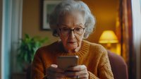 Senior woman in glasses and orange sweater using smartphone at home, warm evening lighting creates cozy atmosphere. Elderly person learning modern technology.