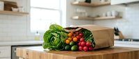 Fresh organic vegetables spilling from brown paper grocery bag on wooden kitchen counter in modern home interior for healthy cooking and meal preparation.