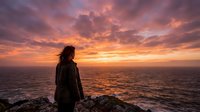 Woman with long dark hair, wearing casual jacket, standing on rocky cliff overlooking ocean at dramatic sunset with golden and purple sky, contemplative mood, travel and nature concept.