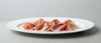 Thinly sliced cured ham arranged on a white oval plate against a light gray background, showcasing delicate pink meat texture.