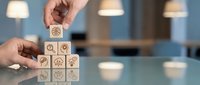 Hands stacking wooden cubes with business and technology icons on a glass table, symbolizing strategy, innovation, and teamwork in a modern office.